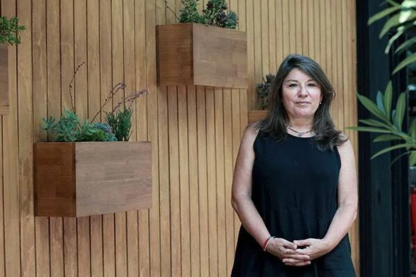 Mujer con vestido negro posa frente a una pared de madera con macetas colgantes que contienen plantas suculentas.