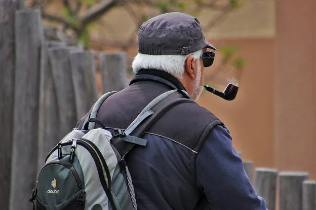 Hombre mayor con cabello blanco, gorra, mochila y pipa en la boca, visto de espaldas en un entorno exterior.