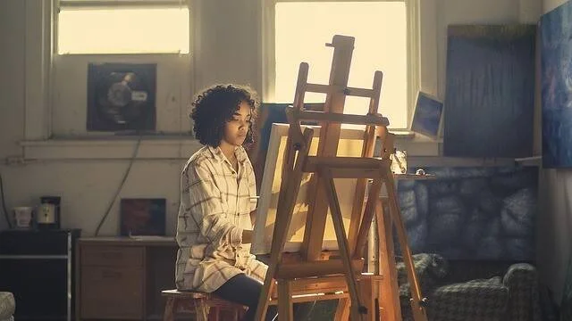 Mujer joven de cabello rizado sentada frente a un caballete de madera, pintando en un estudio de arte iluminado.