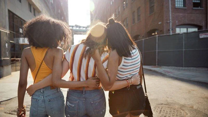 Tres jóvenes caminan de espaldas abrazadas por una calle urbana soleada.
