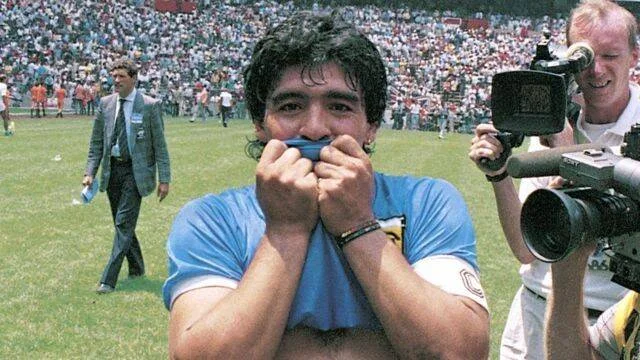 Jugador de fútbol con camiseta celeste besa su camiseta en el campo, rodeado de camarógrafos y público en las tribunas.
