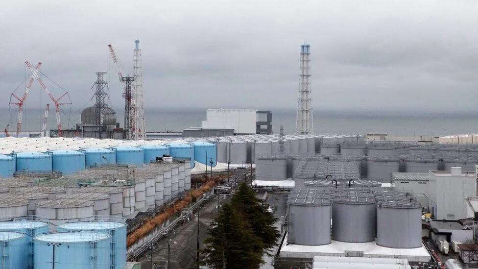 Cientos de tanques de almacenamiento de agua azules y grises en una planta nuclear frente al océano bajo cielo nublado.