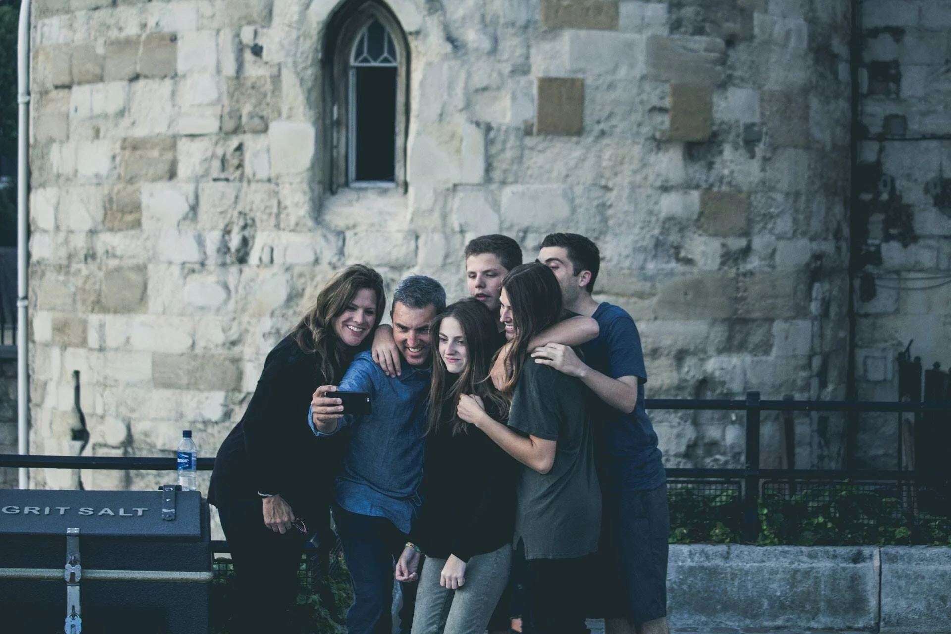 Grupo de seis personas, adultos y jóvenes, se abrazan y miran un teléfono para hacerse una selfie frente a un muro de piedra histórico.