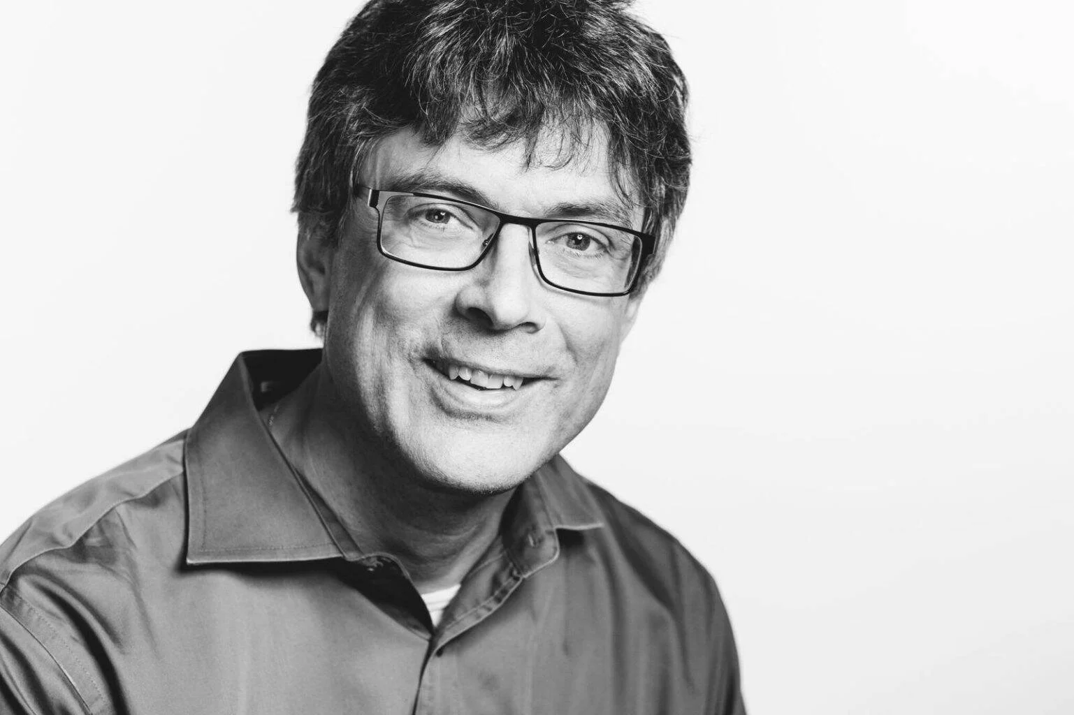 Hombre de mediana edad con gafas rectangulares y camisa, sonriendo en retrato profesional en blanco y negro.