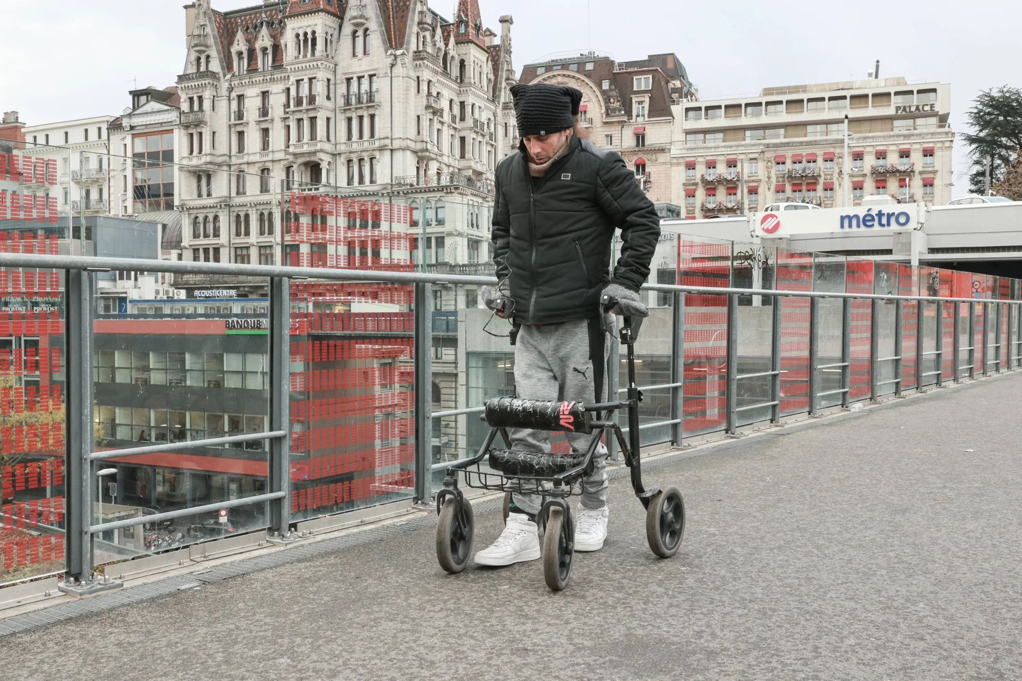 Hombre joven con andador de ruedas camina por un puente urbano cerca de una estación de metro en Lausana, Suiza.