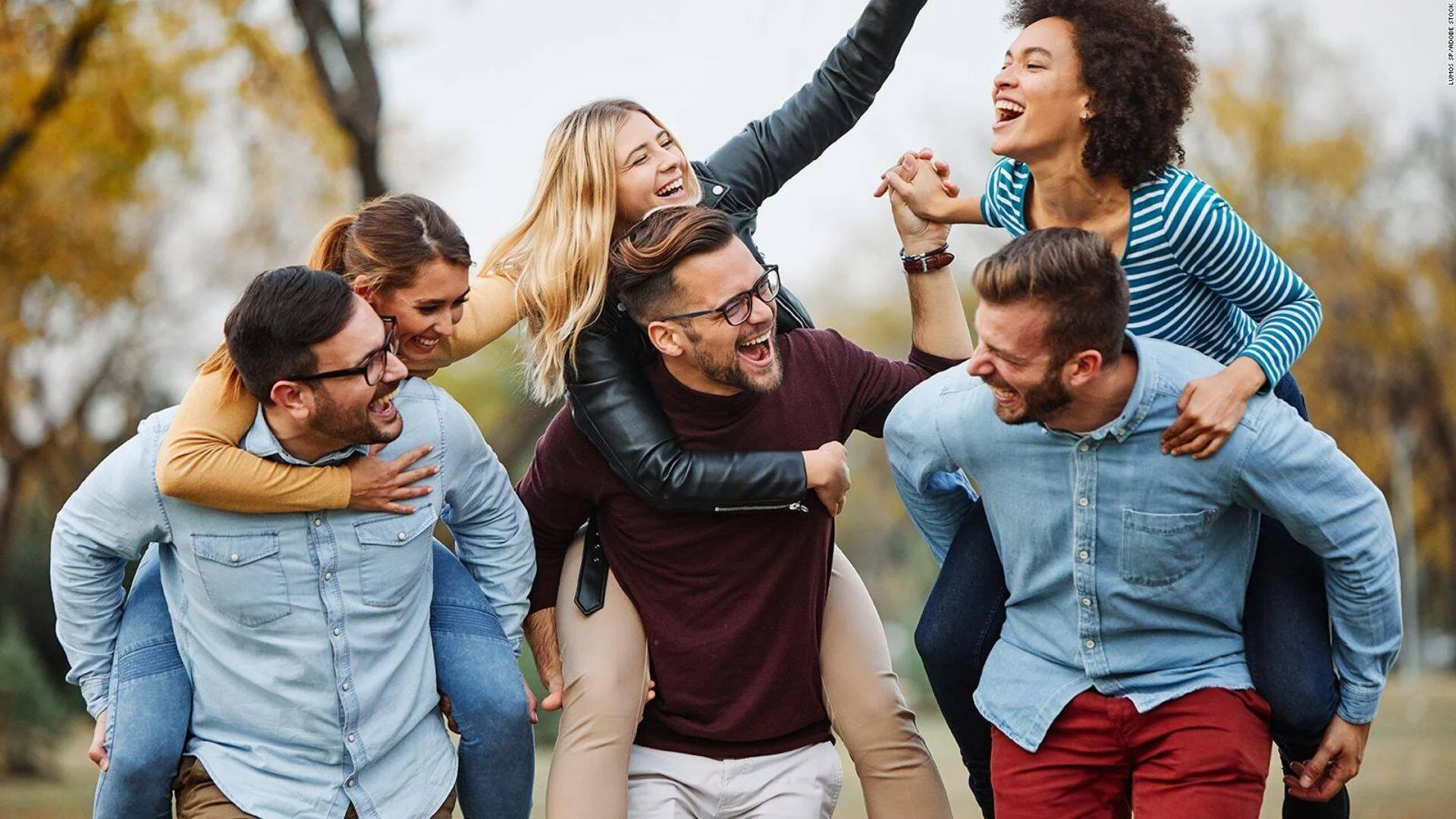 Seis jóvenes amigos ríen y se divierten en un parque, con las chicas subidas a los hombros de los chicos.