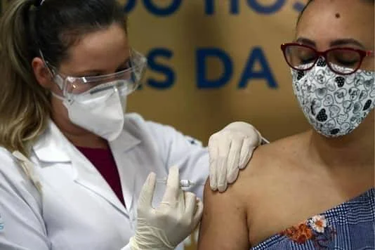 Una enfermera con bata blanca, gafas protectoras y mascarilla aplica una vacuna en el brazo de una mujer con tapabocas.