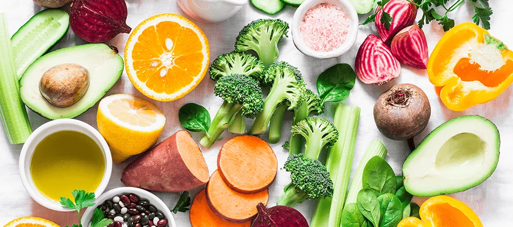 Surtido de alimentos saludables: aguacate, naranja, brócoli, batata, remolacha, pimiento, apio y legumbres sobre superficie blanca.