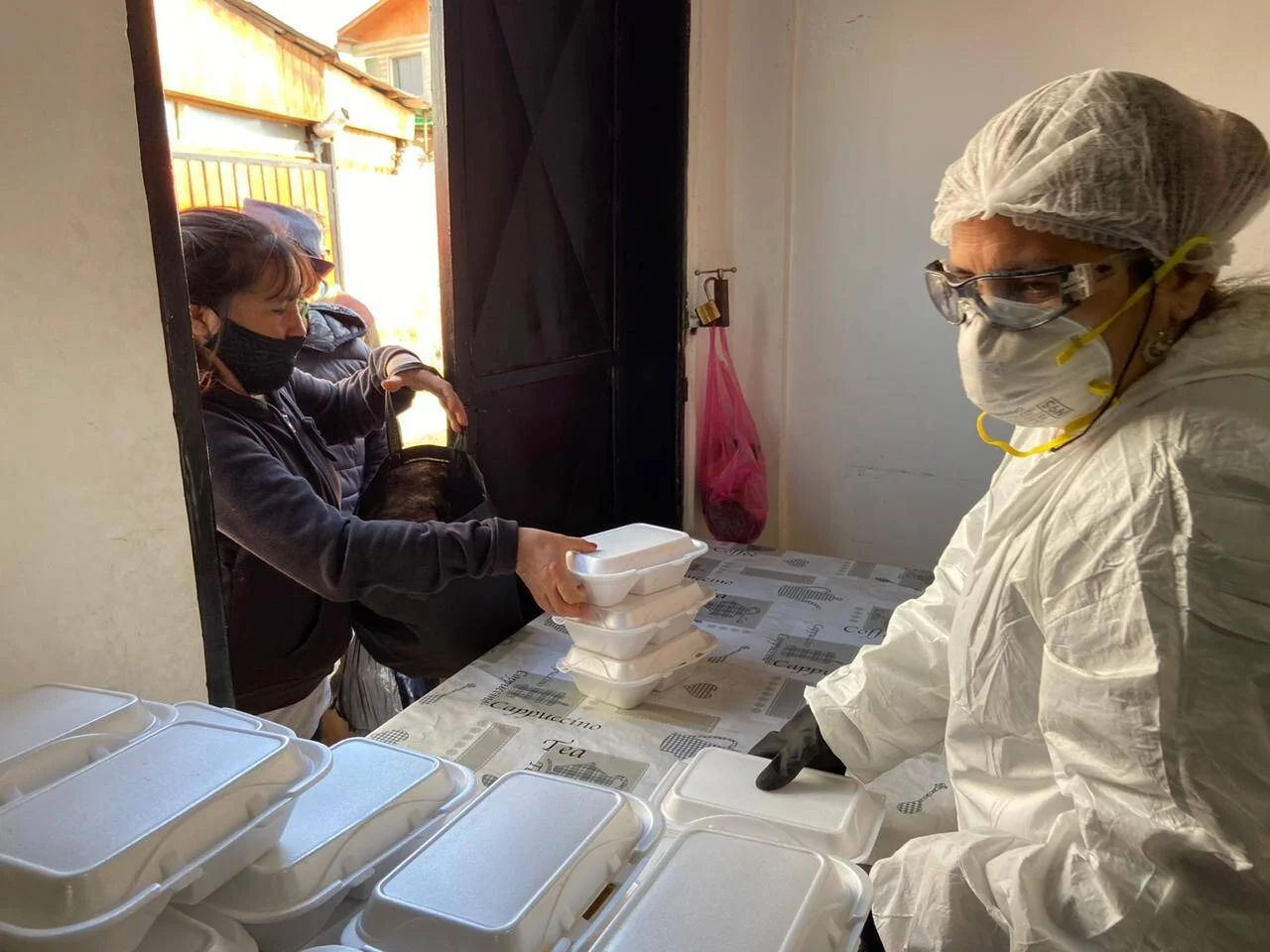 Un voluntario con traje protector, mascarilla y gafas entrega recipientes de espuma a una persona con barbijo en la puerta de un comedor comunitario.