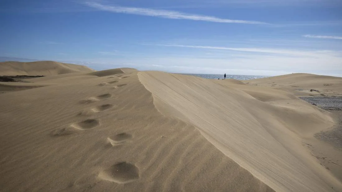 Extensas dunas de arena con huellas de pasos y una persona solitaria en la cresta frente al océano.