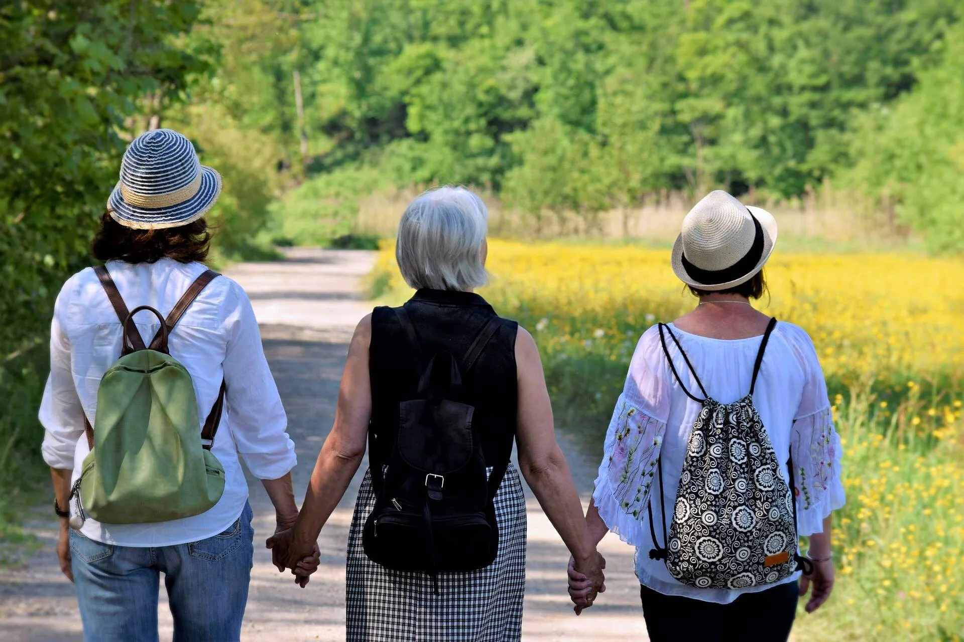 Tres mujeres con mochilas caminan tomadas de la mano por un sendero rodeado de árboles y flores amarillas.