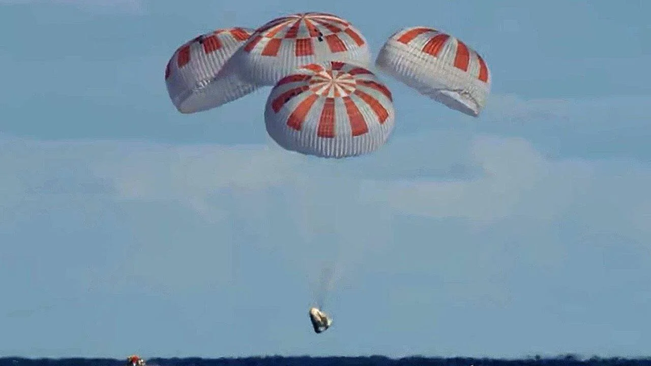 Cápsula espacial suspendida de cuatro paracaídas rojos y blancos desciende sobre el océano durante su amerizaje.