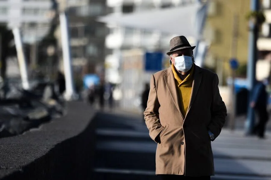 Hombre mayor con sombrero, abrigo y mascarilla blanca camina por una calle urbana en un día soleado.