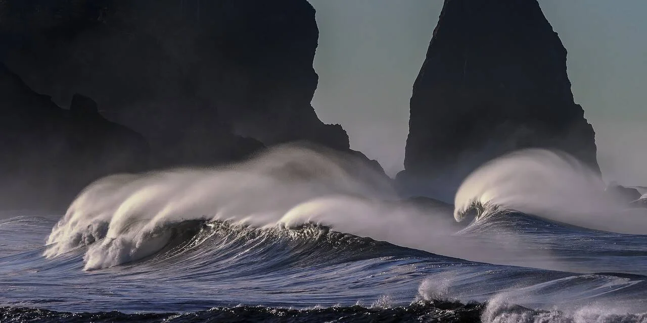 Olas de gran altura rompen con fuerza frente a imponentes formaciones rocosas negras en una costa tempestuosa.