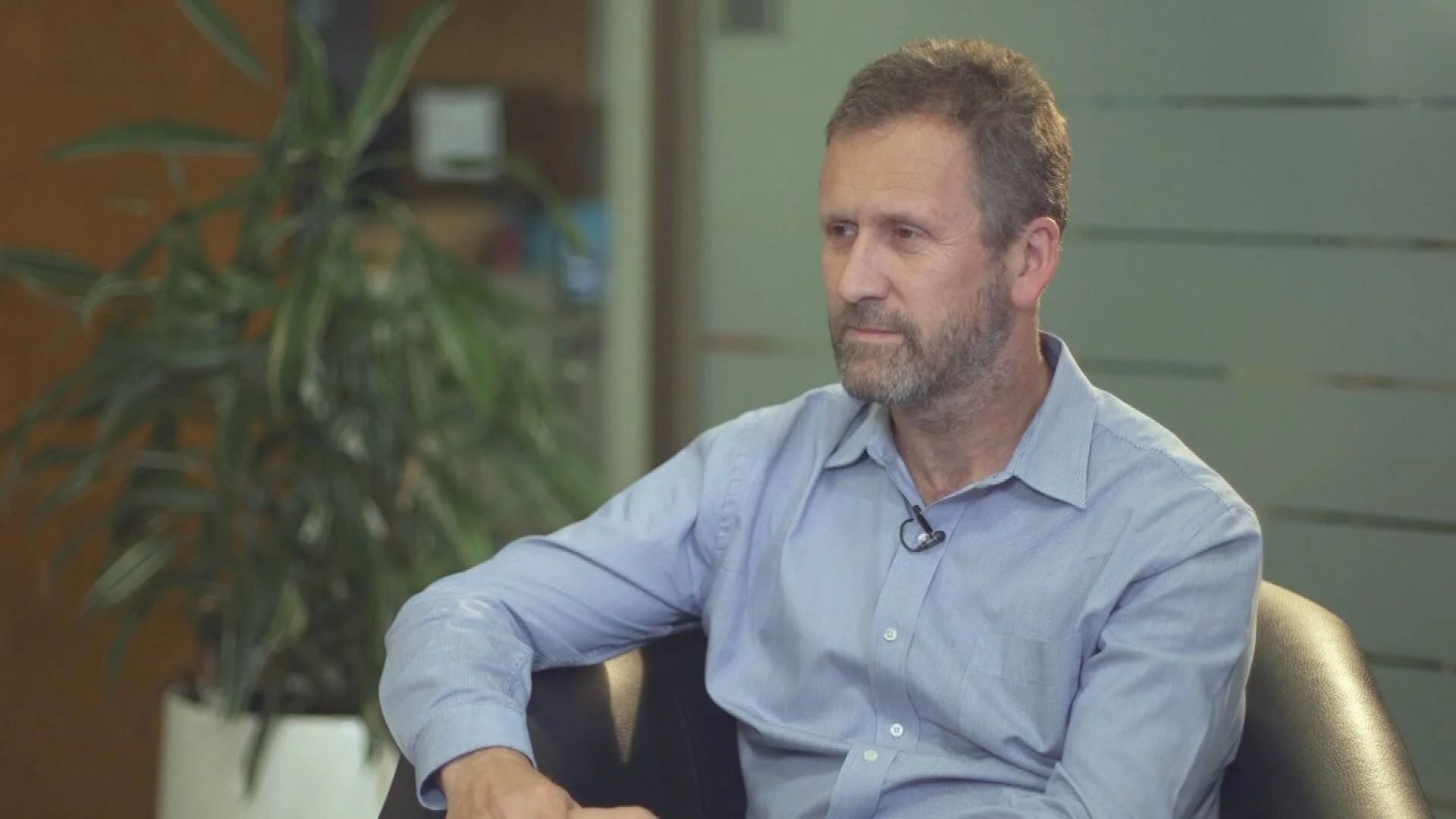 Hombre de mediana edad con camisa azul y micrófono de solapa sentado durante una entrevista en una oficina.