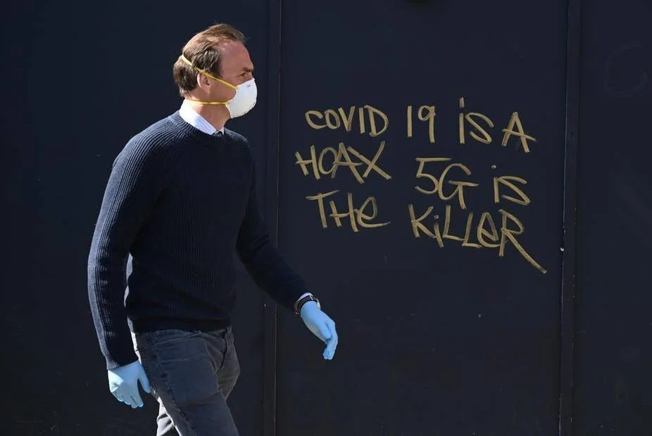 Un hombre con mascarilla y guantes camina frente a un grafiti que dice 'COVID 19 is a hoax, 5G is the killer'.