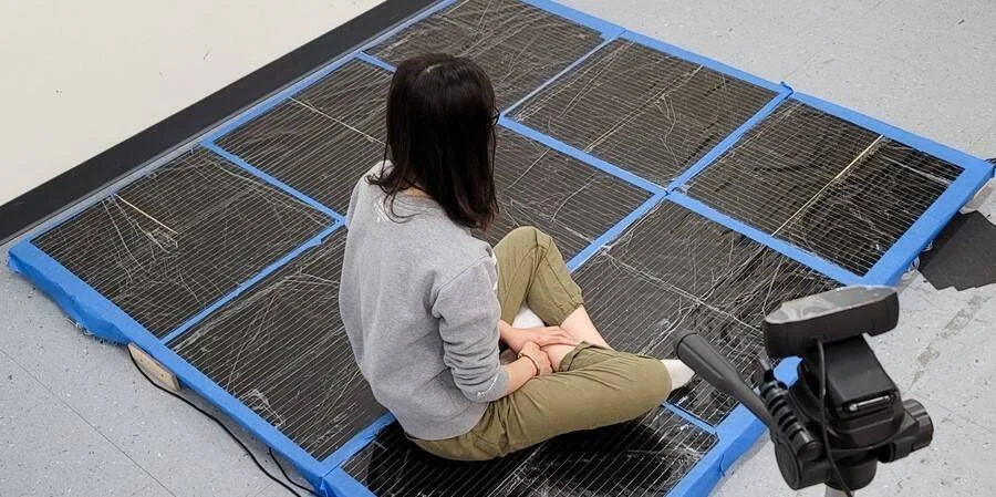 Una mujer sentada en el suelo sobre un conjunto de paneles solares delimitados con cinta azul, frente a una cámara en trípode.