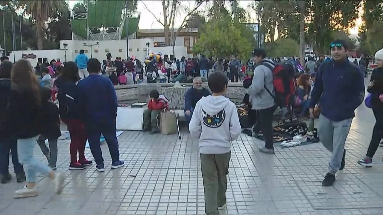 Numerosas personas, incluidos niños y familias, recorren una plaza pública animada con puestos y un escenario al fondo.