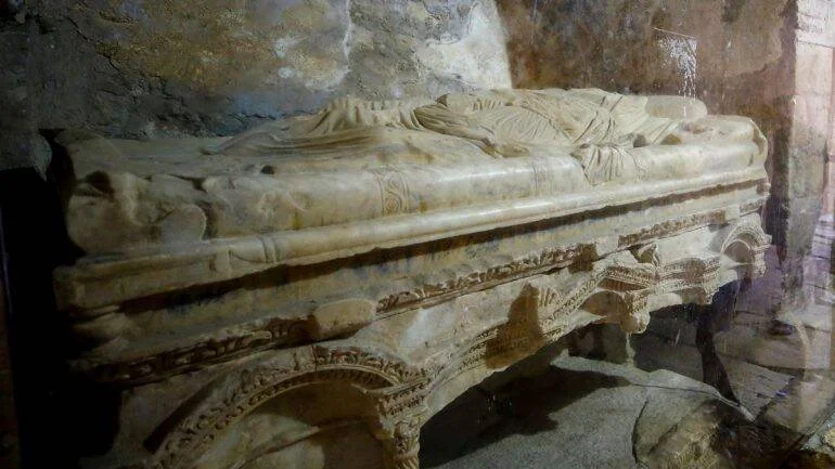 Sarcófago de mármol con escultura funeraria de figura humana yacente, decorado con relieves tallados, en cripta de piedra.