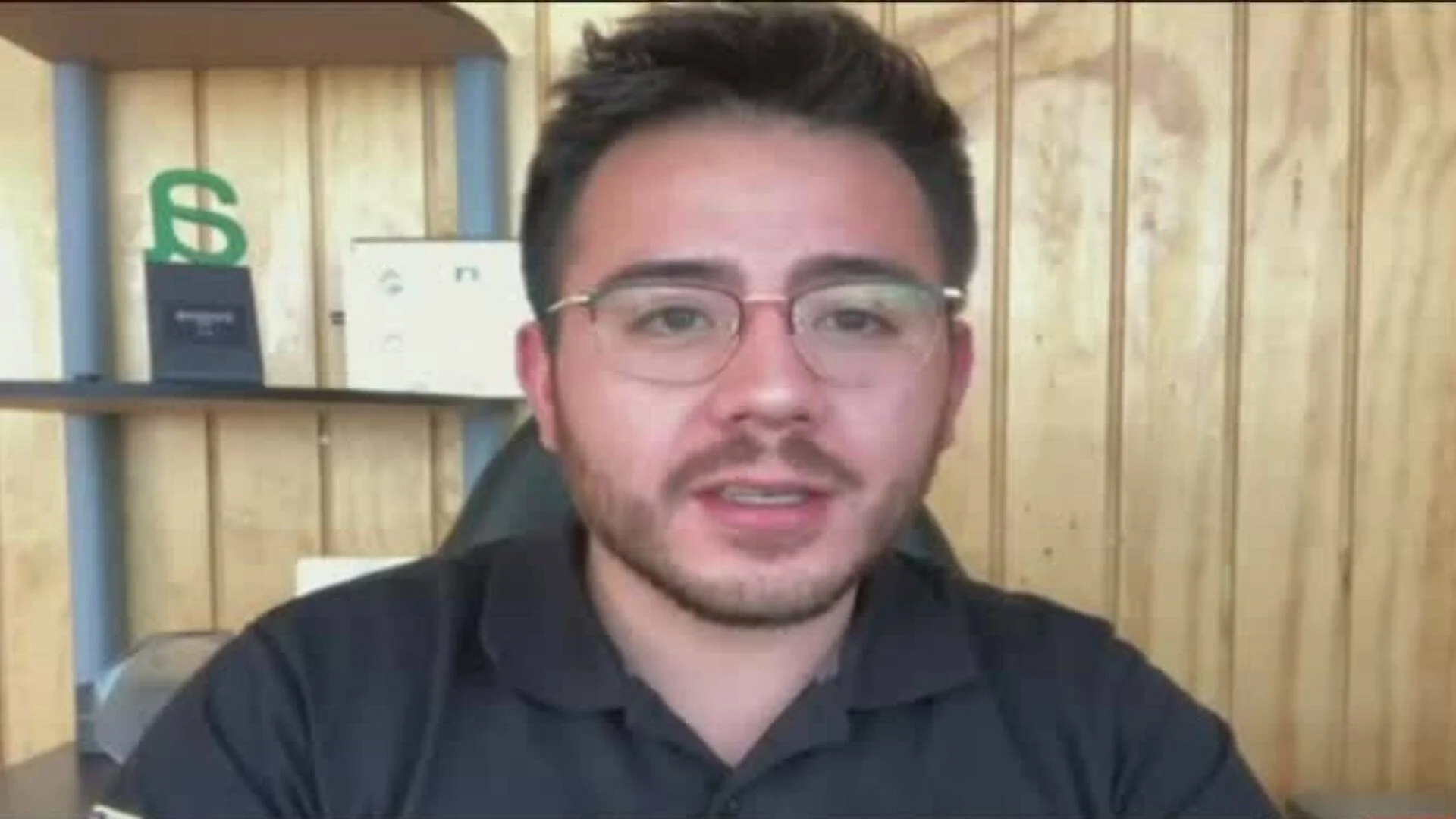 Hombre joven con lentes y remera negra habla frente a cámara en una oficina con pared de madera.