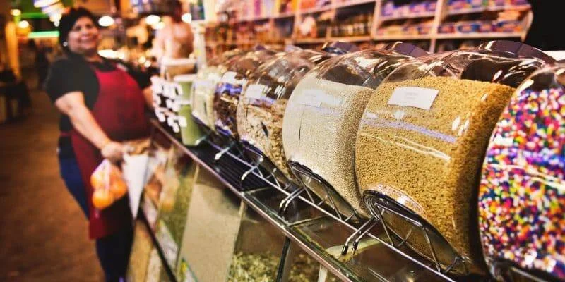 Mujer con delantal rojo sonríe junto a dispensadores de granos, semillas y dulces de colores en un mercado.