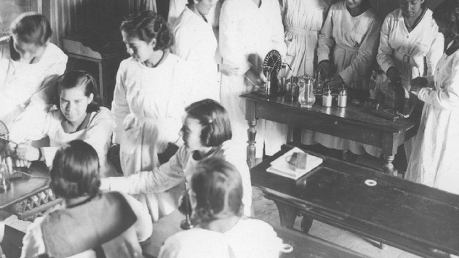 Fotografía histórica en blanco y negro de alumnas con guardapolvo blanco realizando prácticas en un laboratorio escolar.