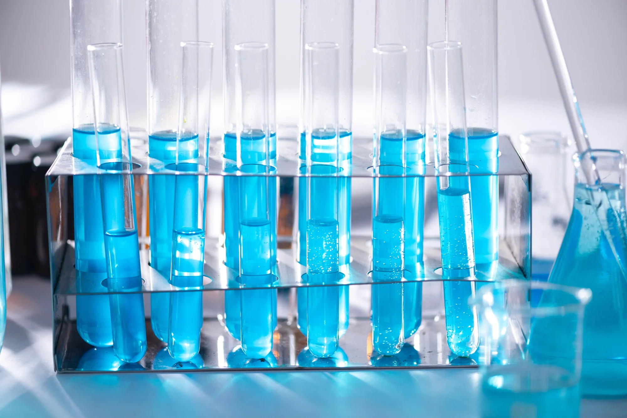 Gradilla metálica con tubos de ensayo llenos de líquido azul junto a un matraz y vaso de precipitados en laboratorio.