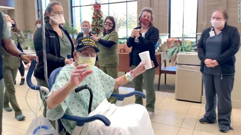 Anciano veterano de la Segunda Guerra Mundial en silla de ruedas es despedido por personal médico con mascarillas en el vestíbulo de un hospital.