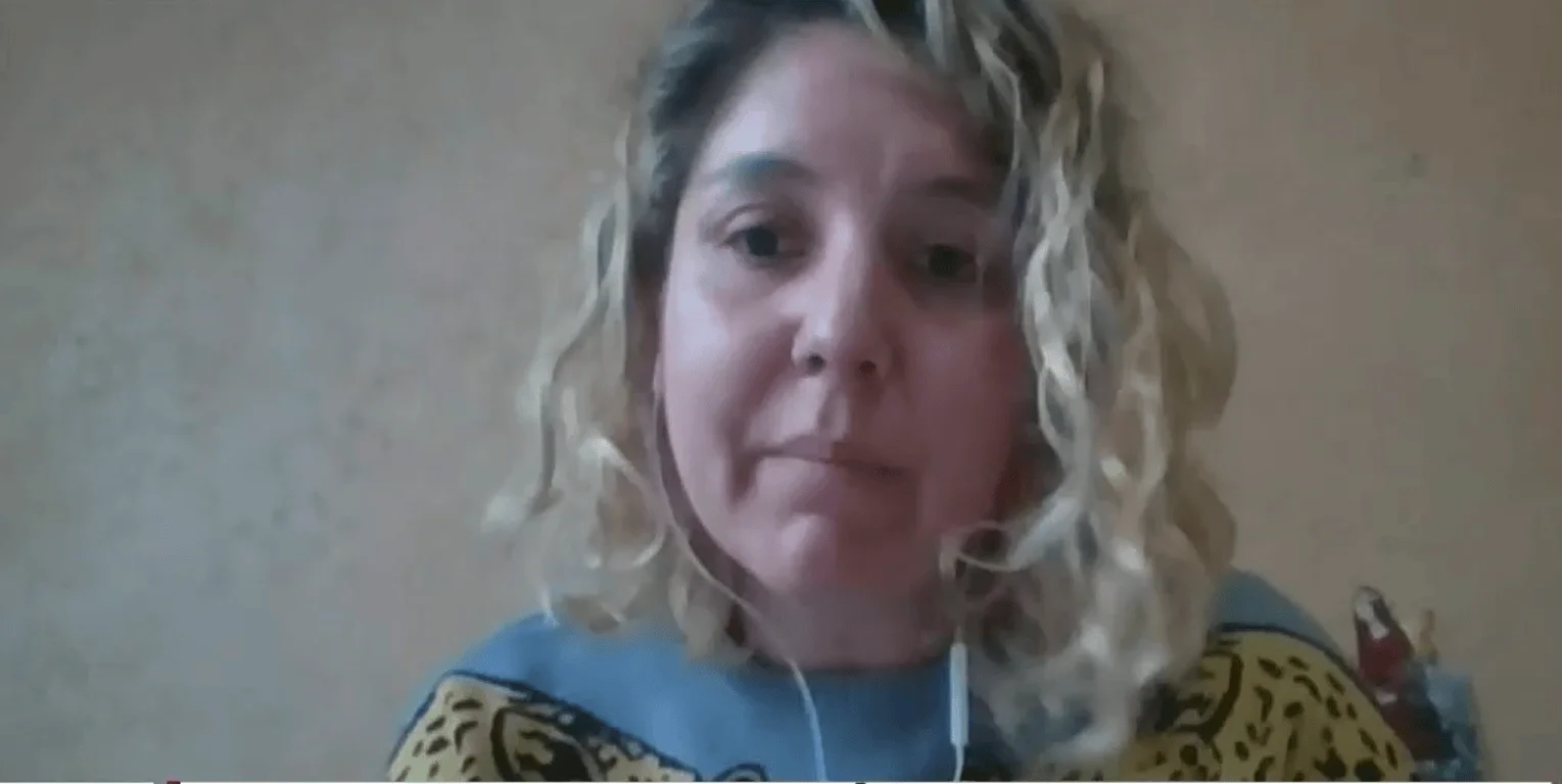Mujer de cabello rizado rubio con auriculares participa en una videollamada frente a una pared beige.