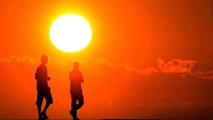 Dos personas caminan en silueta frente a un sol intenso y cielo naranja rojizo, evocando calor extremo.