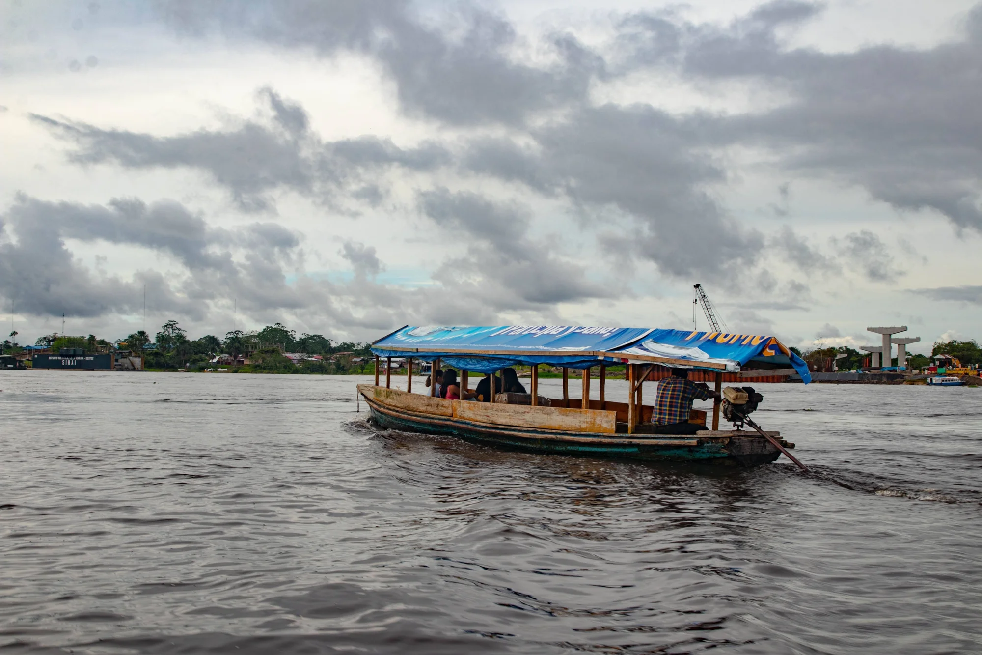 Bote de madera con techo de lona azul transporta pasajeros por un río amazónico bajo cielo nublado, con poblado al fondo.