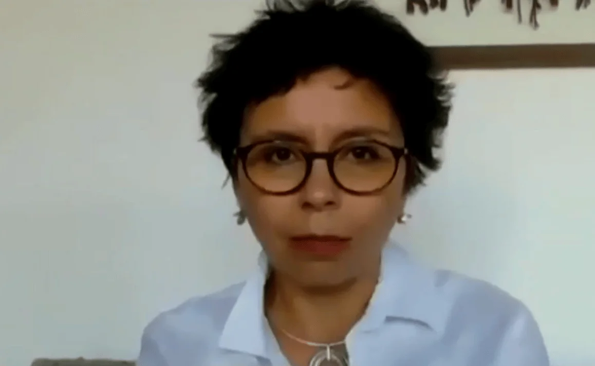 Mujer de cabello corto oscuro y anteojos de carey participa en una videollamada frente a una pared clara.
