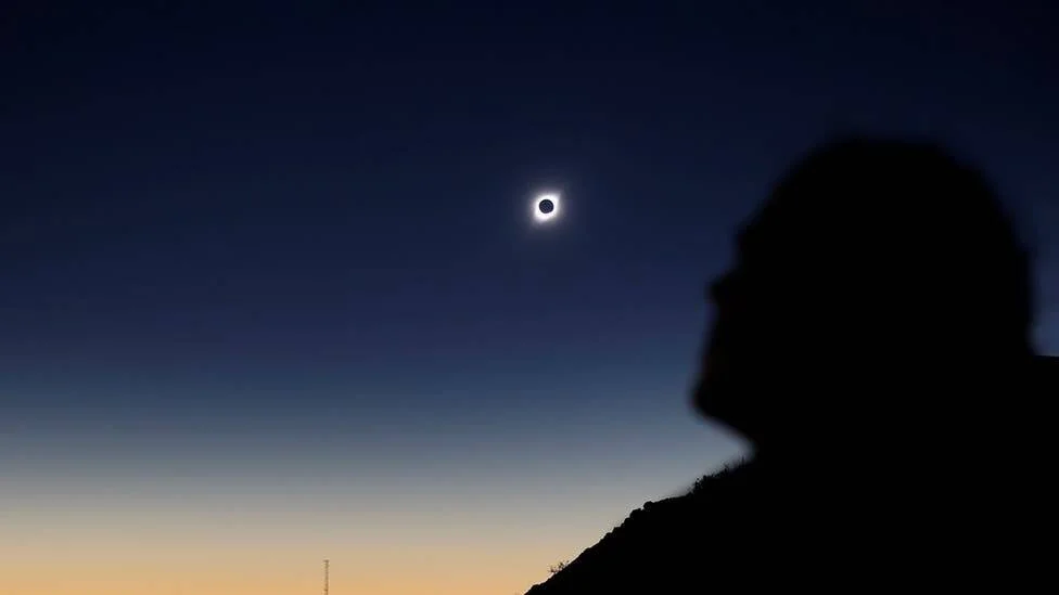 Silueta de una persona contempla un eclipse solar total con la corona visible en un cielo crepuscular azul y naranja.