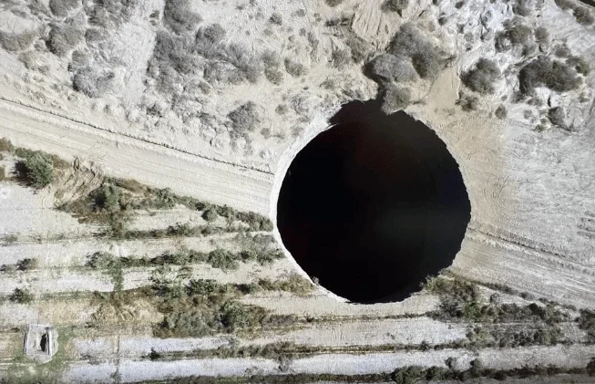 Vista aérea de un enorme socavón circular de fondo oscuro e insondable abierto en un terreno seco y árido.