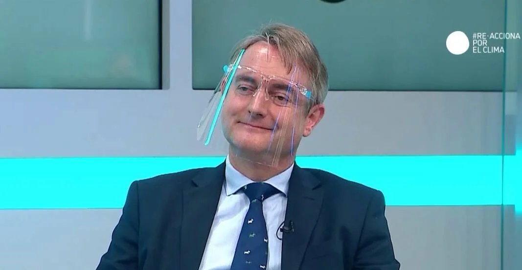Hombre con traje oscuro y pantalla facial protectora sonríe durante una entrevista televisiva sobre el clima.