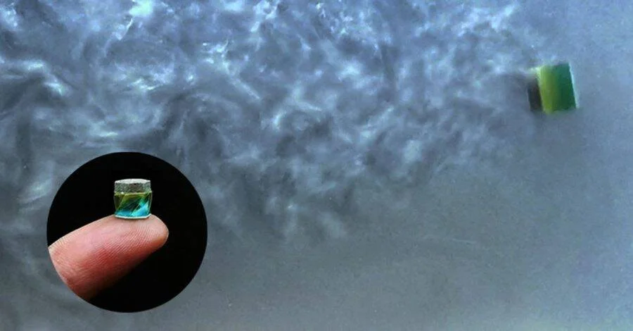 Pequeño robot cilíndrico verde y azul sobre agua, con recuadro que muestra su tamaño real sobre la punta de un dedo.