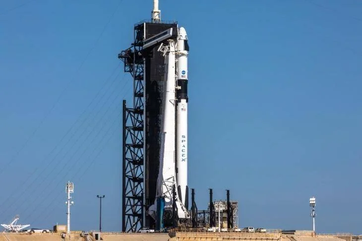Cohete Falcon 9 de SpaceX con cápsula Crew Dragon erguido en la plataforma de lanzamiento bajo un cielo azul despejado.