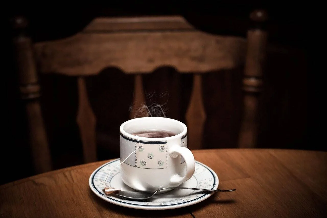 Taza blanca con té caliente humeante, platillo y cucharita sobre una mesa de madera rústica con silla al fondo.