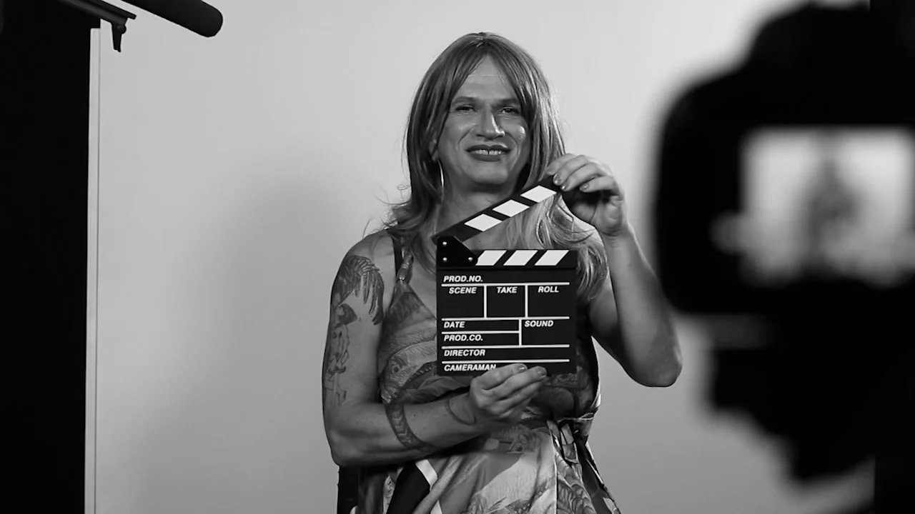 Persona con tatuajes y cabello largo sostiene una claqueta de cine frente a una cámara durante una producción audiovisual.