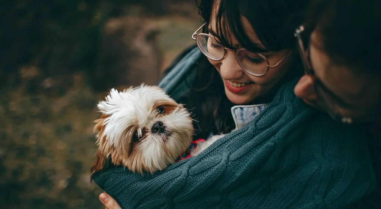 Mujer joven con anteojos sonríe mientras sostiene un pequeño perro Shih Tzu entre sus brazos al aire libre.