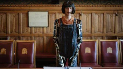 Robot humanoide con apariencia femenina y brazos mecánicos visibles, de pie en una sala de aspecto parlamentario con sillas rojas.