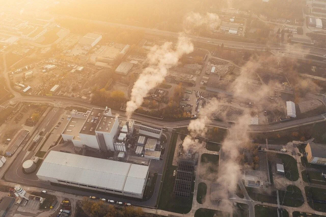Complejo industrial urbano visto desde el aire con columnas de humo emanando de sus chimeneas bajo luz dorada.