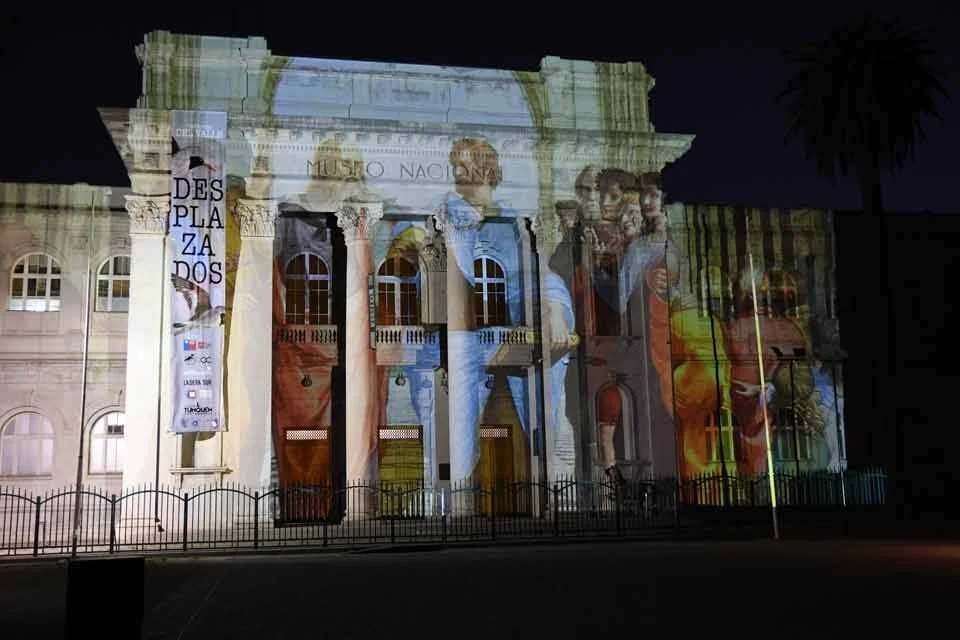 Fachada del Museo Nacional iluminada de noche con una proyección de figuras clásicas y el texto 'Desplazados'.