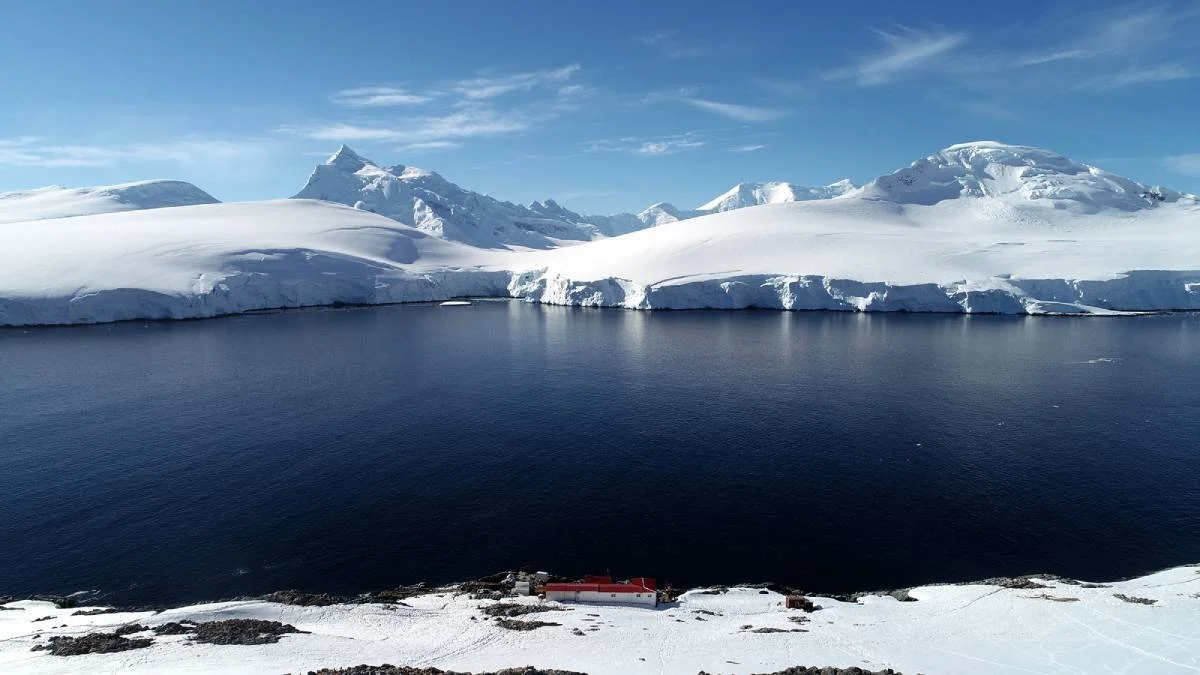 Pequeña base antártica de techo rojo a orillas de una bahía polar rodeada de glaciares y montañas nevadas bajo cielo despejado.