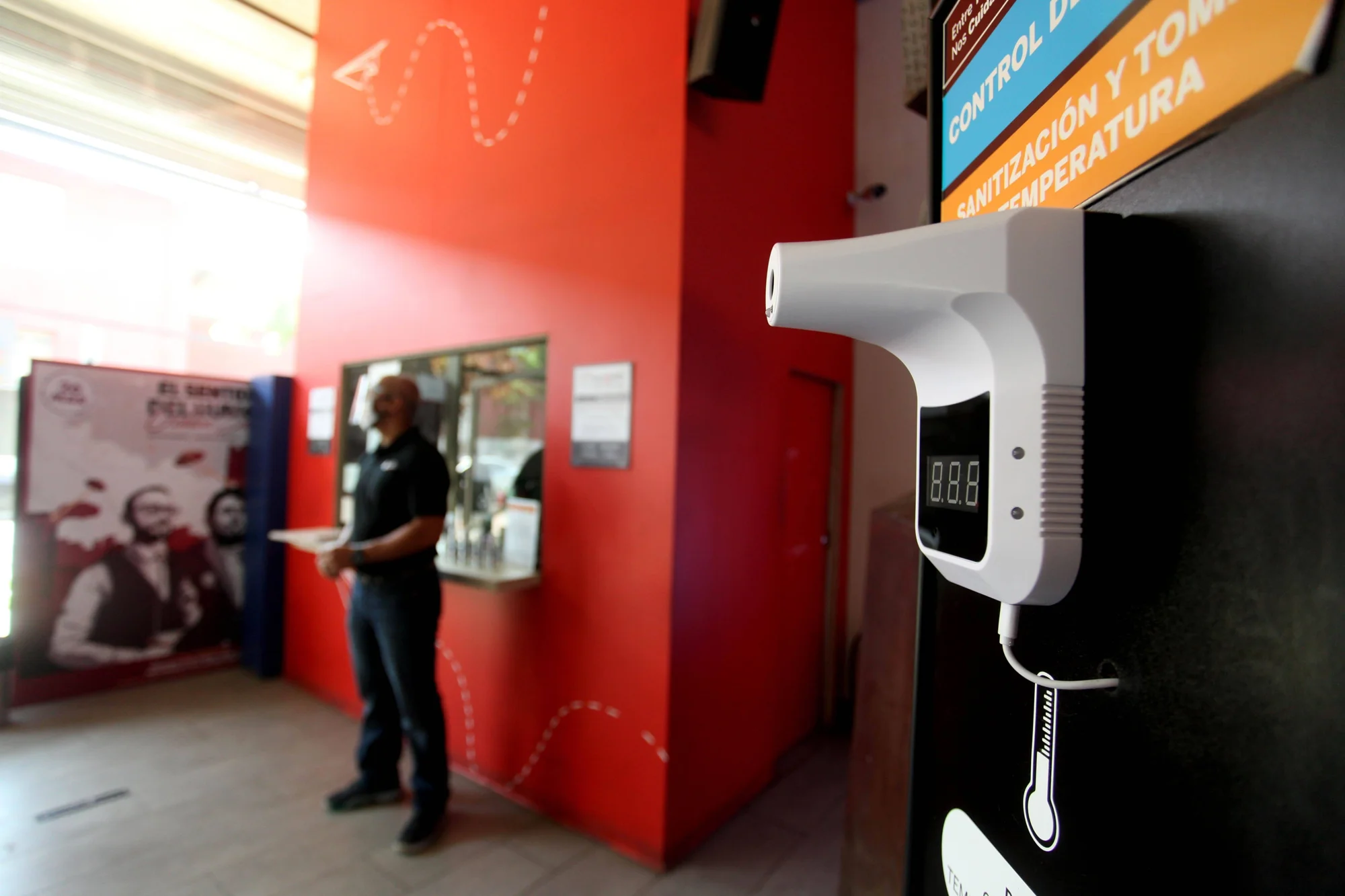 Termómetro infrarrojo sin contacto instalado en la entrada de un local con cartel de control de temperatura y sanitización.