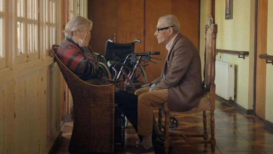 Dos personas mayores sentadas frente a frente en un pasillo con silla de ruedas al fondo; ella cubre su rostro con la mano.