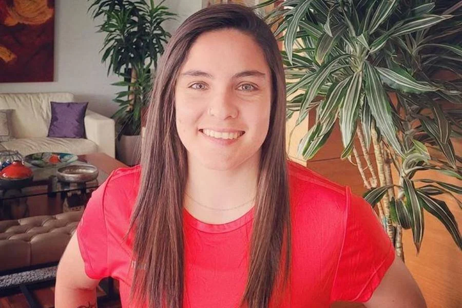 Mujer joven de cabello largo castaño, vestida con remera roja, sonríe a cámara en interior de una vivienda.