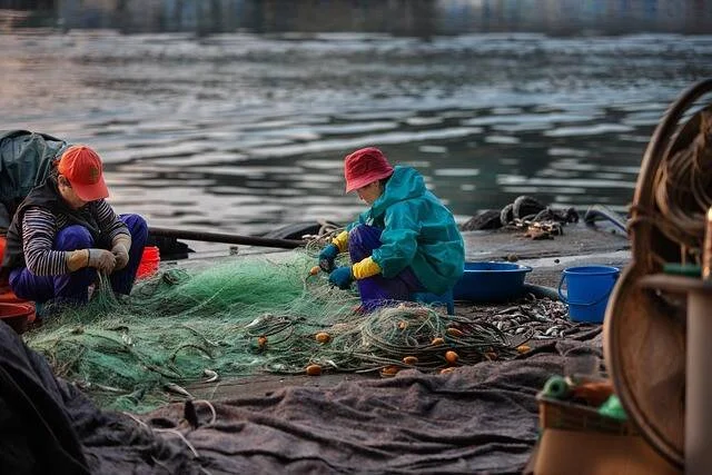 Dos mujeres con gorras y ropa de trabajo remiendan redes de pesca verdes en un muelle junto al agua.