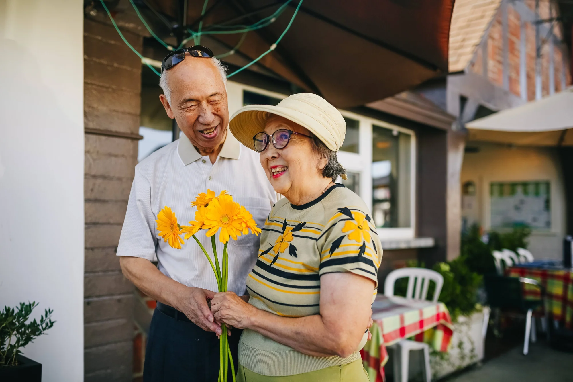 Pareja de adultos mayores sonriendo juntos mientras sostienen un ramo de gerberas amarillas frente a su hogar.