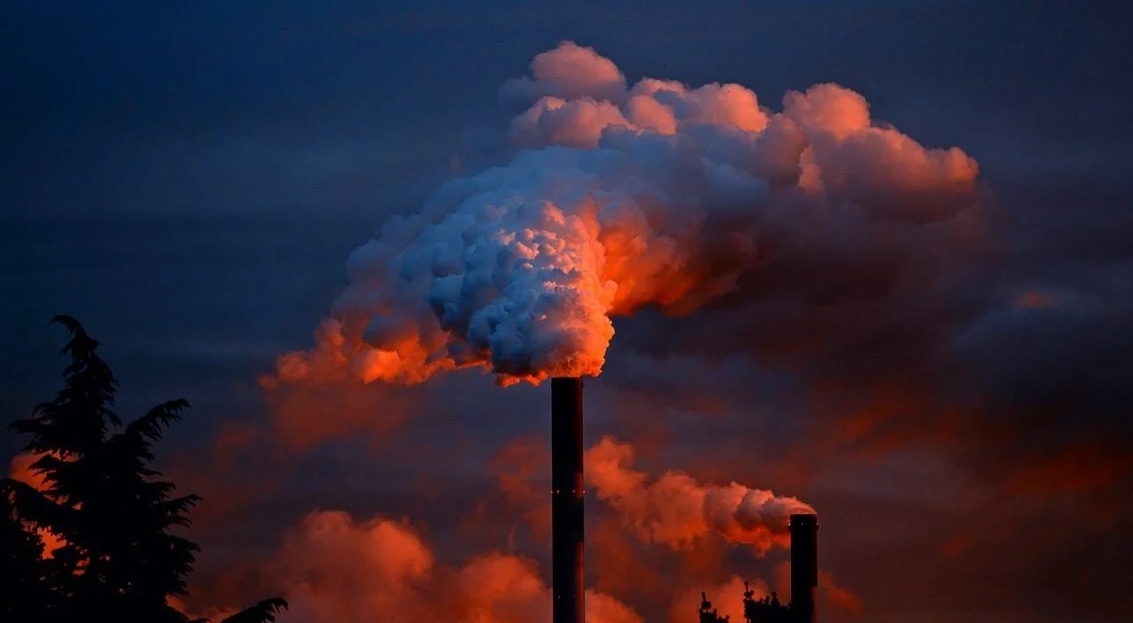 Chimeneas de una planta industrial expulsan grandes nubes de humo iluminadas por la luz rojiza del atardecer.